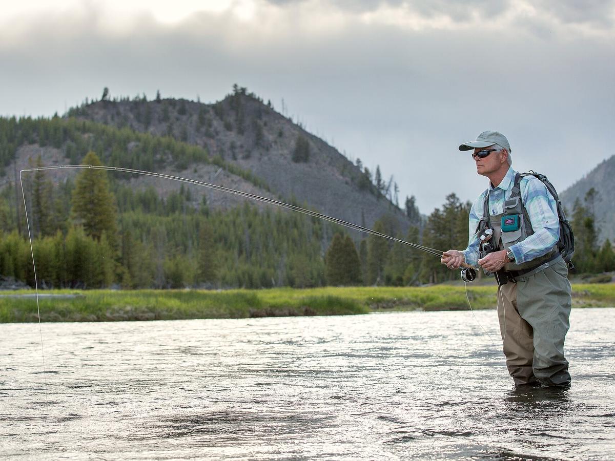 A fly fisherman standing knee-deep in a clear mountain river, pulling in his line